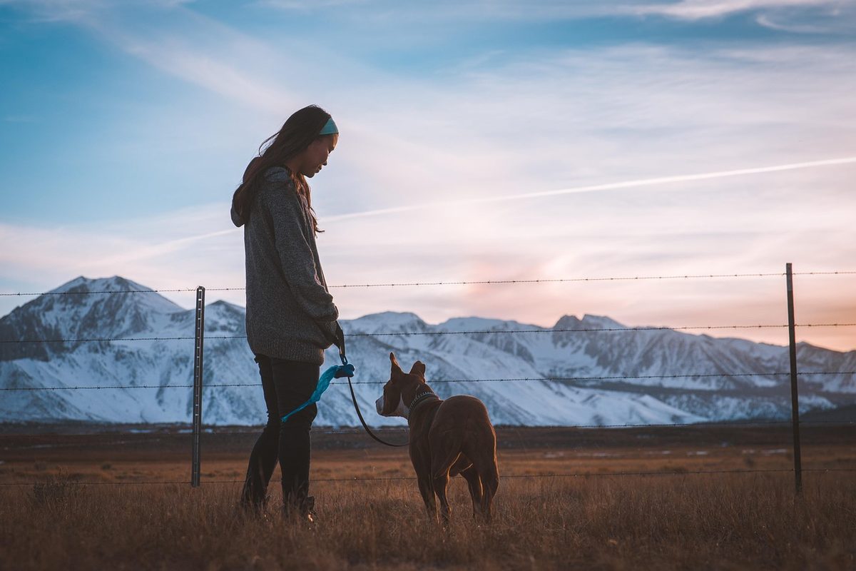 Frau mit Stirnband steht mit einem braunen Hund an der Leine auf einer Wiese vor schneebedeckten Bergen